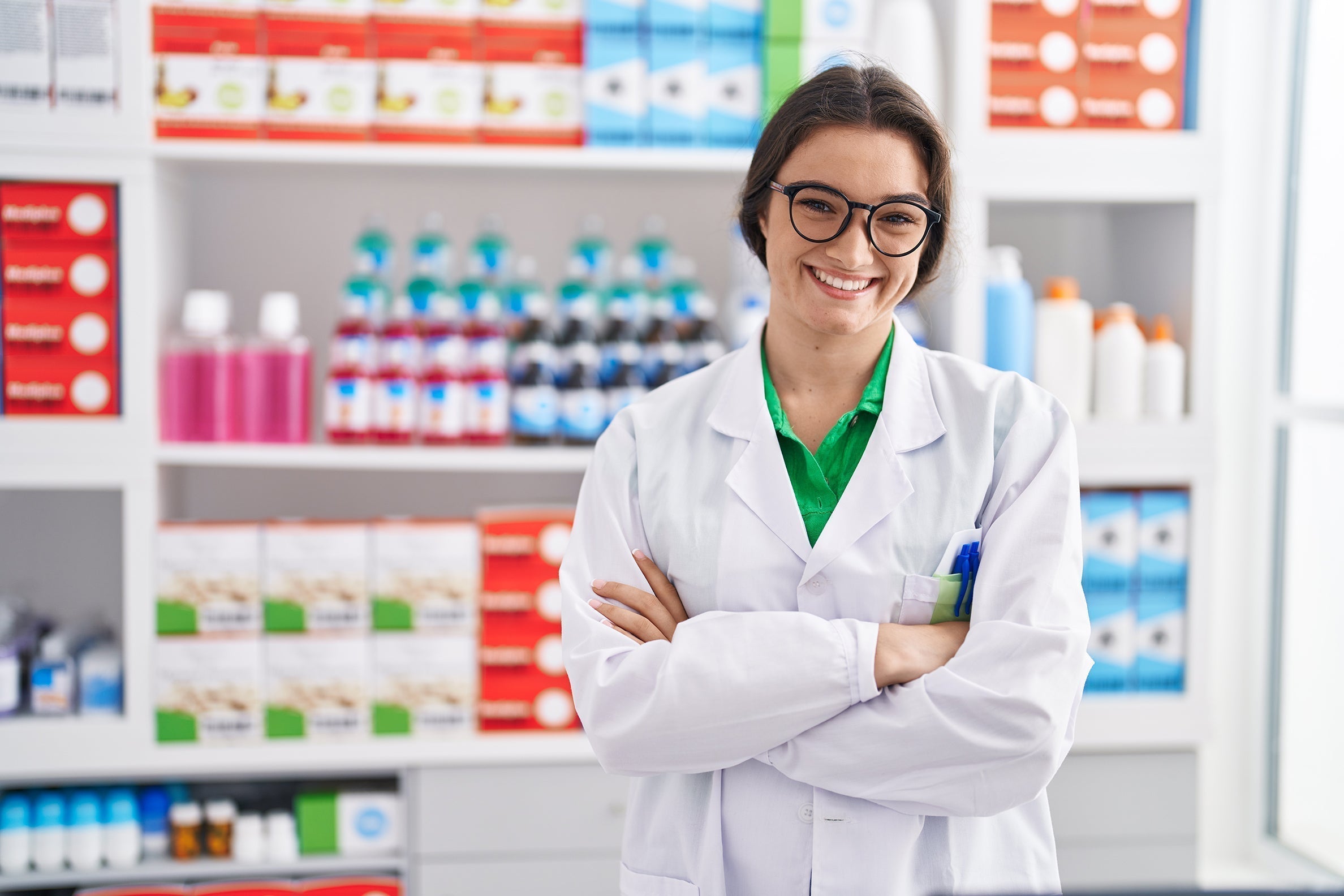 Farmacéutica joven sonriendo con los brazos cruzados en una farmacia moderna, transmitiendo confianza y profesionalidad.